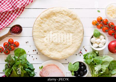 Pizza-Teig und Zutaten auf weißem Hintergrund aus Holz. Cherry-Tomaten, Basilikum, Schinken, Mozzarella, Parmezan, schwarze Oliven, Gewürze und grüner Mangold Stockfoto