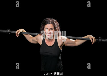 Eine weibliche Gewichtheber entspannt mit Arme ausgestreckt über eine Langhantel, die über ihre Schultern.  Sie für die Kamera lächelt und ist Kreuz beleuchtet auf eine b Stockfoto