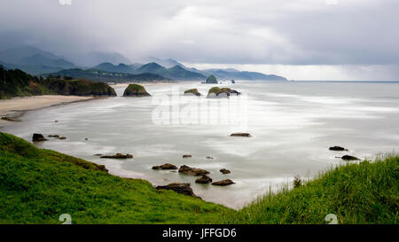 Langzeitbelichtung von Cannon Beach in Oregon, USA mit Felsvorsprüngen in den Ozean und die Berge im Hintergrund an einem bewölkten Himmel-Tag Stockfoto
