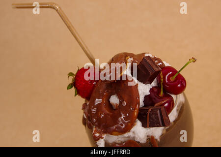 Nahaufnahme von Schokolade nachsichtig extreme Milchshake mit Brownie Kuchen, Erdbeeren, Kirschen und ein Kunststoff Stroh mit Milchschaum oben in einem weichen braun Stockfoto
