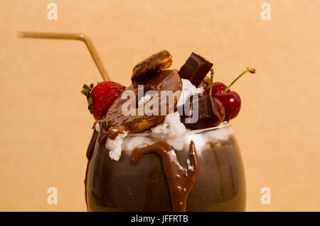 Nahaufnahme von Schokolade nachsichtig extreme Milchshake mit Brownie Kuchen, Erdbeeren, Kirschen und ein Kunststoff Stroh mit Milchschaum oben in einem weichen braun Stockfoto