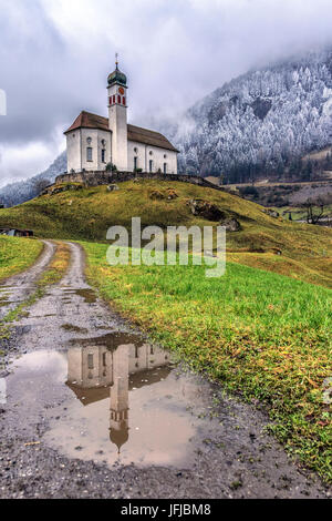 Die Kirche von Wassen spiegelt sich in einer Pfütze nach einem Winter Schneefall, Andermatt, Kanton Uri, Schweiz, Europa Stockfoto