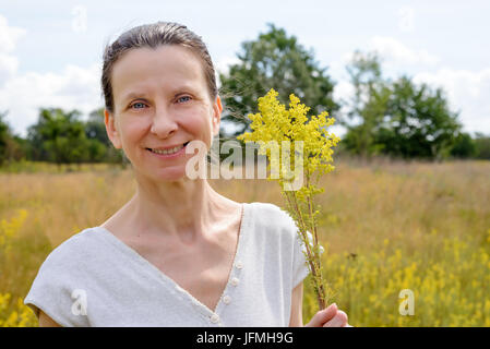 Porträt einer erwachsenen Frau stehend in einer Wiese voller Galium Verum Blumen, auch bekannt als lady's Labkraut oder gelbe Labkraut, mit einem Haufen o Stockfoto