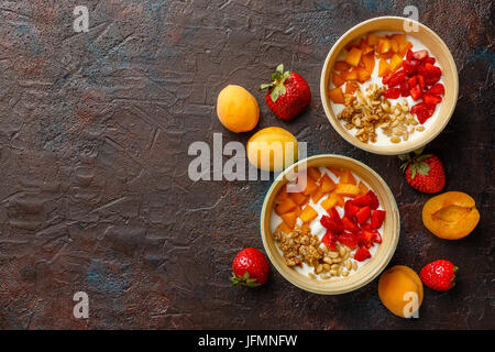 Naturjoghurt mit Stücken von Aprikosen, Erdbeeren, Müsli und Pinienkernen in zwei Schüsseln auf dunklem Hintergrund Broun. Draufsicht mit Platz für text Stockfoto