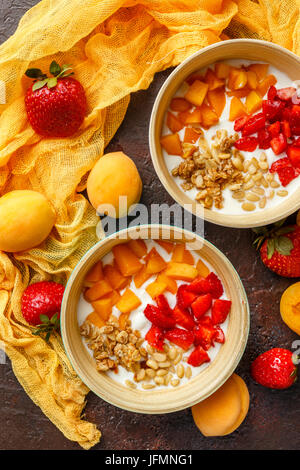 Naturjoghurt mit Stücken von Aprikosen, Erdbeeren, Müsli und Pinienkernen in zwei Schüsseln auf dunklem Hintergrund Broun. Ansicht von oben Stockfoto