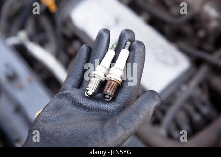 Auto Mechaniker tragen schützende Arbeitshandschuh hält alte und neue Zündkerzen über einem Automotor Stockfoto