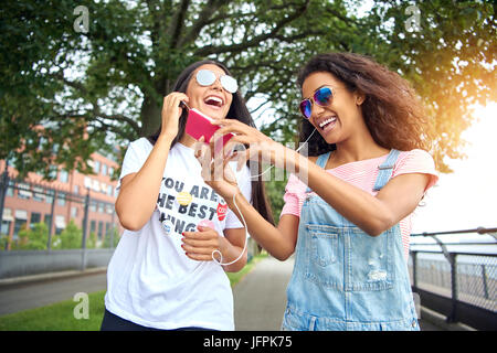 Zwei junge Freunde Lächeln Lachen und zusammen hinunter eine Promenade im Sommer Musik über Kopfhörer hören Stockfoto