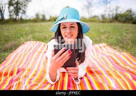 Attraktive lächelnde junge Frau lässig gekleidet verwendet e-Buch/Tablet liegend auf einer bunten Decke im Park. Moderner Lifestyle mit tragbaren mobi Stockfoto