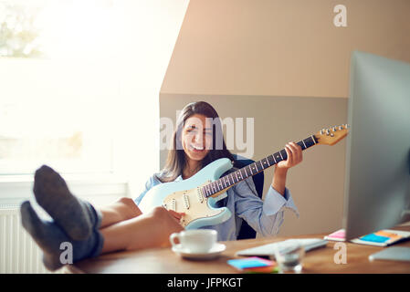Single freudige junge schöne Frau mit Füßen auf den Tisch und spielen E-Gitarre vor Monitor Stockfoto