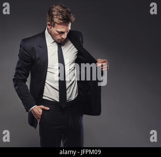 Suave junger Geschäftsmann, Check-out die Jacke von einem stilvollen schwarzen Anzug allein in einem Studio vor einem grauen Hintergrund Stockfoto