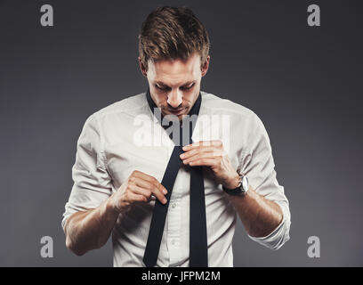 Hübscher junger Geschäftsmann in einem weißen Hemd steht in einem Studio vor einem grauen Hintergrund, seine Krawatte zu binden Stockfoto