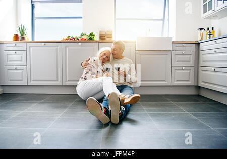 Liebevolle älteres paar lächelnd und zufrieden zusammen auf dem Boden ihrer hellen Küche zu Hause sitzen trinken Rotwein Stockfoto