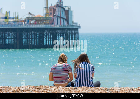 Zwei junge Frauen sitzen am Strand gegenüber der berühmten viktorianischen Struktur, Brighton Palace Pier an einem sonnigen Tag. Stockfoto