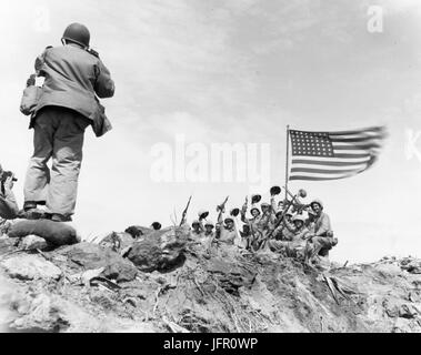 In eine steife Brise auf Mount Suribachi, Vulkan-Berg auf der Insel Iwo Jima, erhöhen die amerikanische Flagge Peitschen gegen den Himmel als Jubel Marine ihre Stimmen und Waffen in den historischen Moment für ein Foto, Iwo Jima, Februar 1945. USMC Foto von Bob Campbell Stockfoto