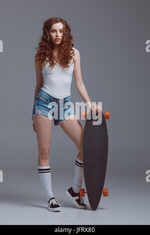 Schöne junge Hipster Frau posiert mit Skateboard isoliert auf grau Stockfoto