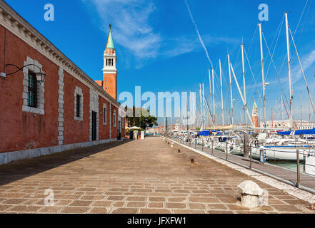 Die Promenade entlang festgemachten Jachten und Glockenturm der Kirche San Giorgio Maggiore auf Hintergrund unter blauem Himmel in Venedig, Italien. Stockfoto