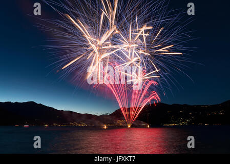 Feuerwerk an der Seepromenade von Luino über den großen See an einem Sommerabend mit blauem Himmel und Berge im Hintergrund Stockfoto