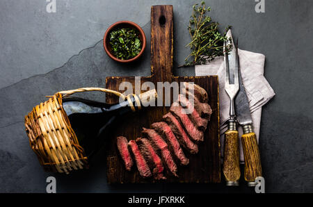 Mittlere seltene Rindersteak auf Woodenboard mit Kräutersauce und Botle Wein, graue Schiefer Hintergrund Stockfoto