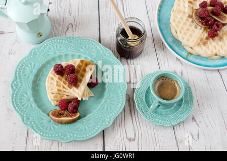 Weiche belgische Waffeln mit Himbeeren und Feigen, in Herzform mit Honig auf türkis blaue Platte bedeckt. Einen Kaffee und eine Kaffeemaschine. Rustikale Holz zurück Stockfoto