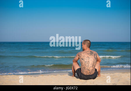 Ein junger Mann mit einem großen Tattoo auf seinem Rücken sitzen am Strand mit Blick aufs Meer. Textfreiraum links Stockfoto