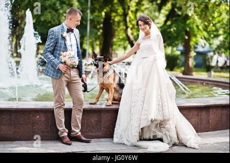 Fabelhafte junge Brautpaar posiert neben einem Brunnen mit dem Hund in den Park. Stockfoto