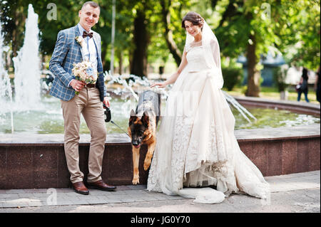 Fabelhafte junge Brautpaar posiert neben einem Brunnen mit dem Hund in den Park. Stockfoto