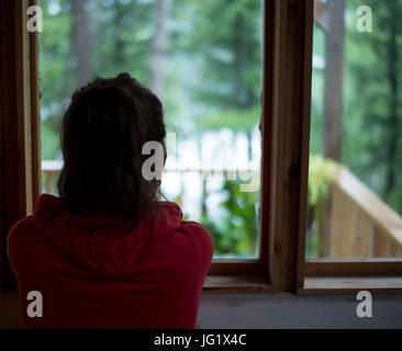 Junge braune behaarte weibliche Blick Fenster Haus am Wald an einem regnerischen Tag. Stockfoto