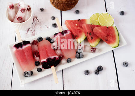 Eis am Stiel mit Wassermelone, Kokosmilch, Kalk, Himbeeren und Heidelbeeren. Eis am Stiel auf einem weißen rechteckigen Plateau, Rechteck-Eis mit Himbeeren. L Stockfoto