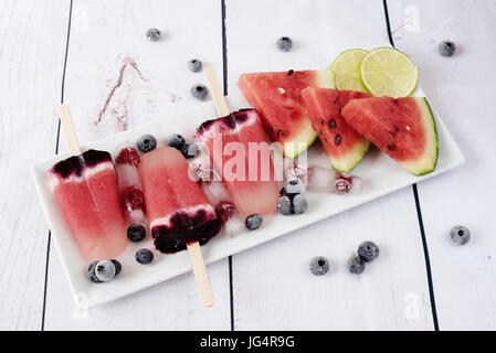 Eis am Stiel mit Wassermelone, Kokosmilch, Kalk, Himbeeren und Heidelbeeren. Eis am Stiel auf einem weißen rechteckigen Plateau, Rechteck-Eis mit Himbeeren. L Stockfoto