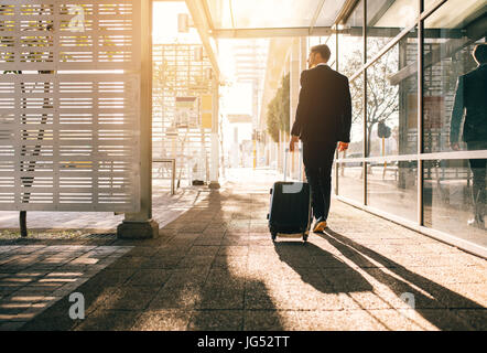 Rückansicht des Kaufmanns zu Fuß mit Tasche außen Flughafen. Junge Geschäftsreisende ziehen Koffer am Stadt Straße. Stockfoto