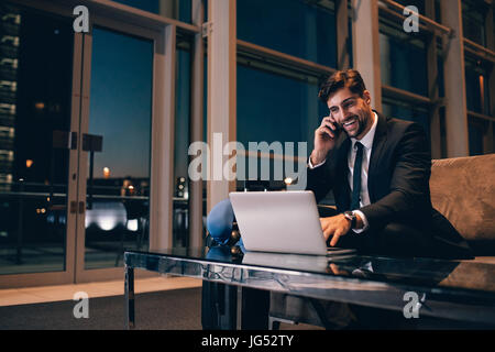 Lächelnd Geschäftsmann mit Laptop in der Wartehalle des Flughafens am Handy sprechen. Gut aussehender Mann im Wartezimmer im Flughafen-terminal. Stockfoto