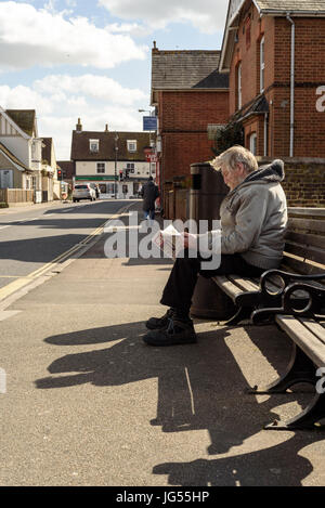 Alter Mann mit einem Bart sitzt auf einer Bank auf der Seite einer Straße lesen der beliebten englischen Zeitung Sun bei Tageslicht Stockfoto