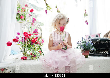 Niedliche kleine Mädchen tolle Kleid sitzt auf der Fensterbank neben die Blumen mit einem Tupil in ihrer Hand. Stockfoto