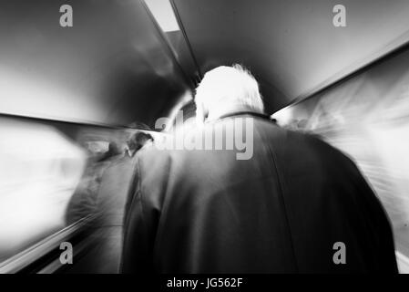 Ein Alter Mann mit weißen Haaren und blauen Mantel auf einer London unterirdische Rolltreppe mit Motion blur in schwarz / weiß Stockfoto