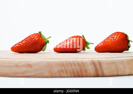 Eine Linie von drei roten Erdbeeren mit Blättern und kurz gestielt intakt, sichern Schuss seitlich auf Augenhöhe, auf ein Schneidbrett aus Holz gegen ein cremefarbenes Stockfoto