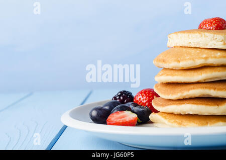 Ein Stapel Frühstück Pfannkuchen belegt mit einer Erdbeere, stehend auf einem weißen Porzellanteller, umgeben von Sommerfrüchten.  Blass blau Holztisch und zurück Stockfoto