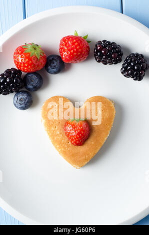 Eine herzförmige Pfannkuchen in der Mitte von einem weißen Teller mit einer Erdbeere zentriert oben drauf.  Darüber, eine Vielzahl von Sommerfrüchten angeordnet sind, einem Stockfoto