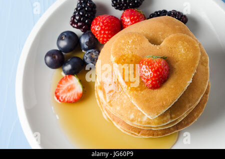 Overhead Schuss aus einem Stapel von Pfannkuchen auf einen weißen Porzellanteller mit Sommerfrüchten, gekrönt mit einem Herz geformten eins und eine Erdbeere. Tropfen mit Ahorn s Stockfoto