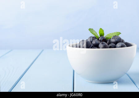 Blick auf Ebene eine weiße Schüssel mit Heidelbeeren garniert mit einem Zweig Minze auf einem Holztisch geplankte hellblau gegen einen hellblauen Hintergrund gemalt. Stockfoto