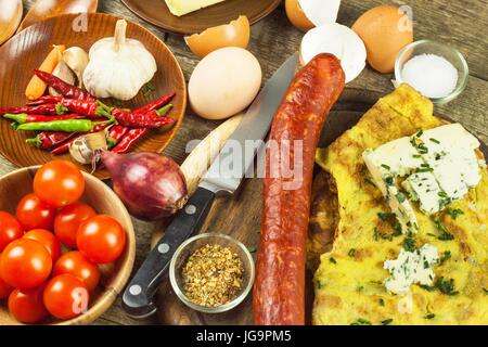 Lecker gefüllte Omelette auf einem Holzbrett. Gebratenes Ei Omelett mit Wurst, Cherry-Tomaten, Chilischoten und Schimmel Käse. Nahrhaftes Frühstück Stockfoto