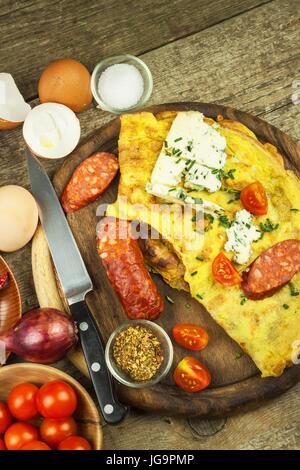 Lecker gefüllte Omelette auf einem Holzbrett. Gebratenes Ei Omelett mit Wurst, Cherry-Tomaten, Chilischoten und Schimmel Käse. Nahrhaftes Frühstück Stockfoto