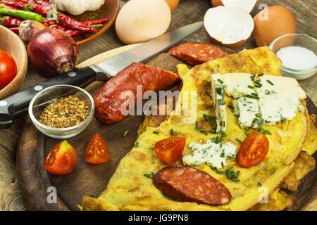 Lecker gefüllte Omelette auf einem Holzbrett. Gebratenes Ei Omelett mit Wurst, Cherry-Tomaten, Chilischoten und Schimmel Käse. Nahrhaftes Frühstück Stockfoto