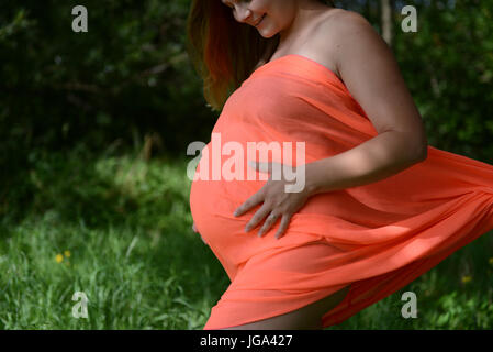 Schwangere junge Frau mit orange Kleid steht in einem park Stockfoto