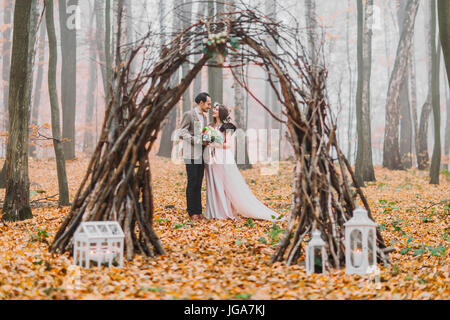 Glücklich Brünette Brautpaar schauen liebevoll einander unter dem kreativen hübsch dekorierte Hochzeit Bogen im herbstlichen Wald Stockfoto