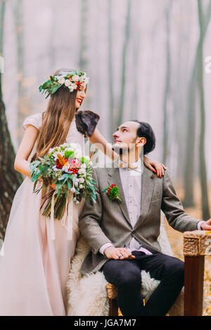 Die fröhliche Braut hält Hochzeit Bouquet von roten Blumen, während streichelte die Schulter des Bräutigams sitzen auf dem Stuhl im Herbst Park. Stockfoto