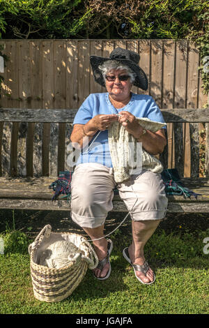 Ein lächelnder Alter/Senioren/ältere Frau/Dame stricken, während auf einer Holzbank sitzen, alleine in einem Garten in der warmen Sonne. England, UK. Stockfoto