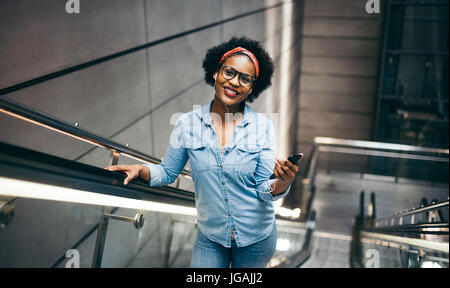 Junge afrikanische Frau lächelnd und hören Sie Musik auf ihr Handy während der Fahrt allein eine Rolltreppe in einem modernen Gebäude Stockfoto