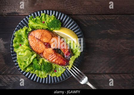 Eine obenliegende Foto ein gegrilltes Lachssteak mit frischen grünen Salat, Brokkoli-Sprossen, eine Scheibe Zitrone und rosa Pfefferkörner, mit einer Gabel auf einem dunklen Stockfoto