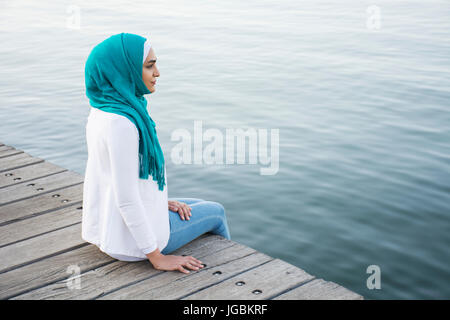 Junge muslimische Frau tragen Hijab saß auf dem Kai am Meer wegschauen Stockfoto