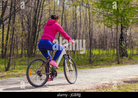 Sportliche junge Frau in einem hellen rosa Jacke und Jeans Fahrradtouren durch den Wald an einem Sommertag. Ansicht von hinten Stockfoto